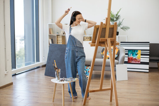 Full length view of young lady in headphones standing barefoot with brush in front of easel with painting. Dancing person drawing inspiration for work from music in wireless device at art space.