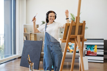 Happy brunette artist in denim jumpsuit handling brush professionally while rocking out to tunes in...
