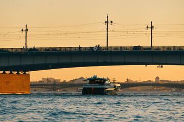 A motor yacht sails under a road bridge at sunset. A luxury boat floats on the river of a European city on a summer evening.