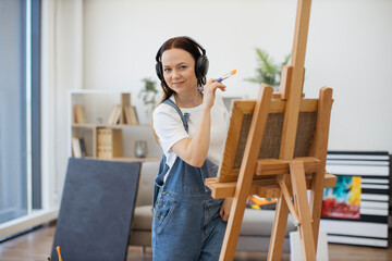Happy brunette artist in denim jumpsuit handling brush professionally while rocking out to tunes in...