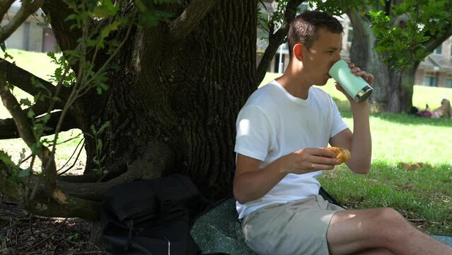 Young man enjoys a delicious lunch sitting on the grass in the park on a sunny summer day.