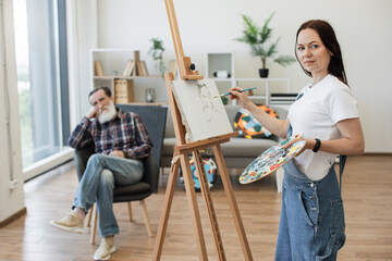 Self-employed female painter in denim overalls depicting sitter head-and-shoulders in modern studio...