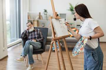 Self-employed female painter in denim overalls depicting sitter head-and-shoulders in modern studio space. Attractive brunette lady sketching out rough ideas of senior man's portrait on canvas.