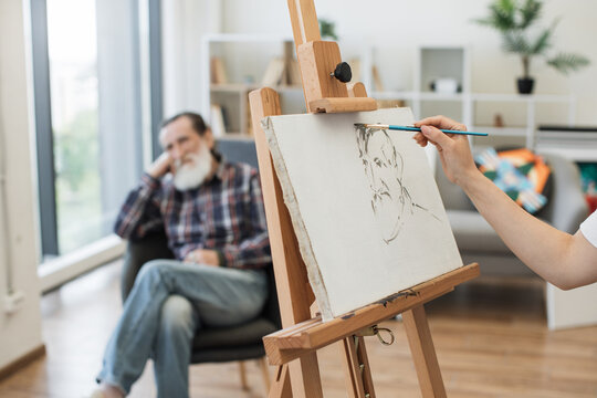 Focus on female hand using paintbrush on cotton canvas supported by easel with male model in background. Fine artist paying attention to details of facial expression while adding interest to work.