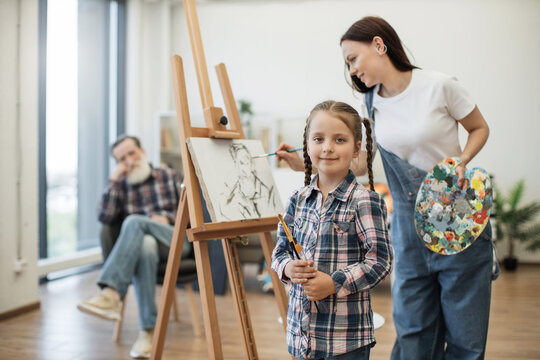 Beautiful girl with paintbrushes looking at camera while young adult drawing from father sitting in background. Talented mother encouraging kid to participate in artistic process at home studio.