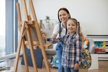 Portrait of proud mother holding daughter's hand with paintbrush while standing near easel with palette. Patient parent training creative tween in sketching on blank canvas at home art workshop.