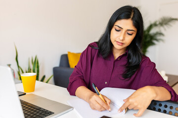 Portrait of young indian woman sitting on table using laptop computer and writing on notebook,...