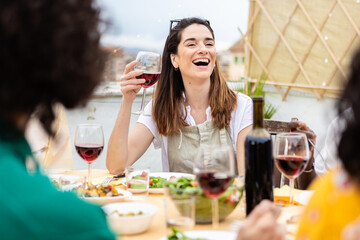 Young group of happy friends enjoying dinner barbecue at home terrace. Smiling people having fun celebrating together eating meat and drinking red wine. Friendship and leisure summer lifestyle concept