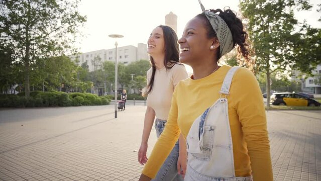Side view of two young beautiful women walking happy street in city on sunny spring day. Two cheerful best friends having fun strolling and talking outdoor. Generation z people laughing together.