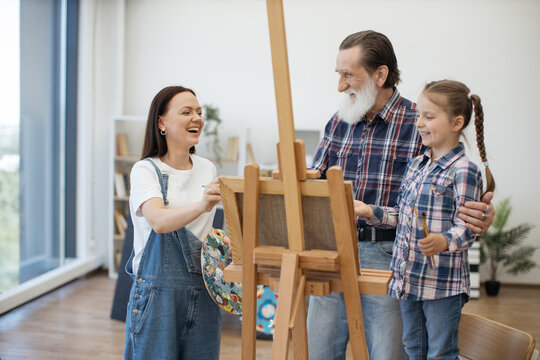 Happy ladies and senior man laughing at image on canvas while standing behind easel in home art space. Smiling mother taking break from serious painting when having fun in studio with dad and kid.