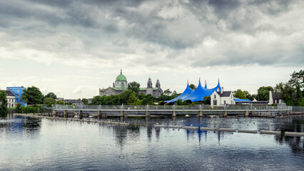 Fototapeta premium Galway city Cathedral building and blue tent by River Corrib. Dark cloudy sky. Popular town landmark. Dark and moody feel.