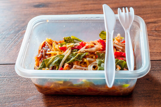 Chicken chow mein noodles in a plastic take away box or container on a wooden table. Quick and tasty Asian style meal for microwave. Lunch in an office concept. Food on a go. High quality ready meal.