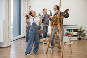 Excited senior man and young females with devices and paintbrushes moving to beat of songs behind...