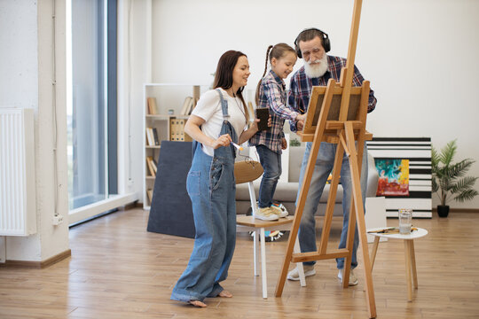Young barefoot woman with earbuds and mobile watching small girl applying paint on cloth near grandpa with headphones. Creative family practising fine art with musical accompaniment in apartment.