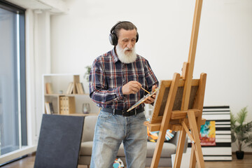 Portrait of smiling pensioner creating artwork while listening to songs via wireless device in room interior. Passionate painter in cozy clothes living full life of inspiration in retirement.