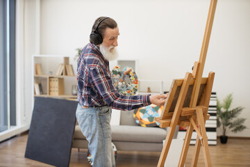 Portrait of smiling pensioner creating artwork while listening to songs via wireless device in room interior. Passionate painter in cozy clothes living full life of inspiration in retirement.