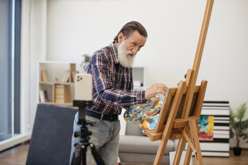Fototapeta na wymiar Casually dressed grey-bearded artist recording video using smartphone on tripod while painting portrait on easel. Concept of older generation using modern technology.