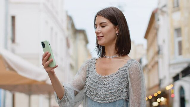Pretty young woman blogger taking selfie on smartphone, communicating video call online with subscribers, recording stories for social media vlog outdoors. Girl walking in urban european city street