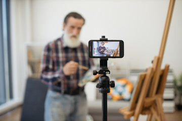 Carefree elder man having online drawing class and using cellphone set on tripod for video conference. Blurred senior gentleman giving advice to young students while standing near easel with palette.