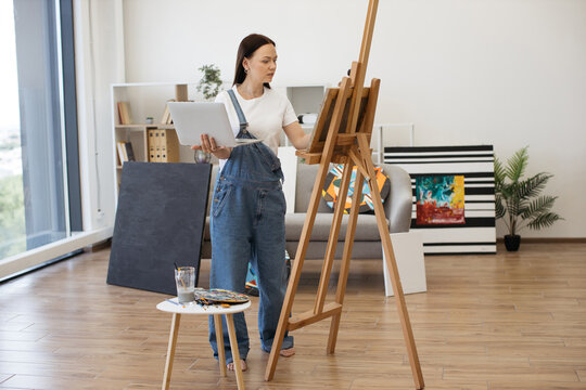 Female professional painter having video conference using portable computer in comfy flat with amazing panoramic view. Attractive female in casual outfit having online art class with beginners.