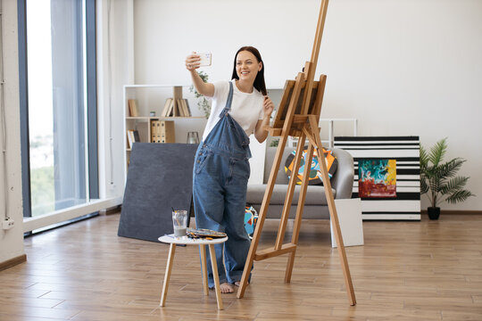 Young beautiful female smiling while making selfie on personal gadget in stylish apartment with large panoramic windows. Creative artist posing near big wooden easel and small table with paints.