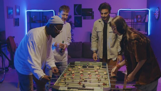 Medium full shot of group of students energetically playing table football at home party, Caucasian girl winning over African American guy, cheering and doing high-fives with friend