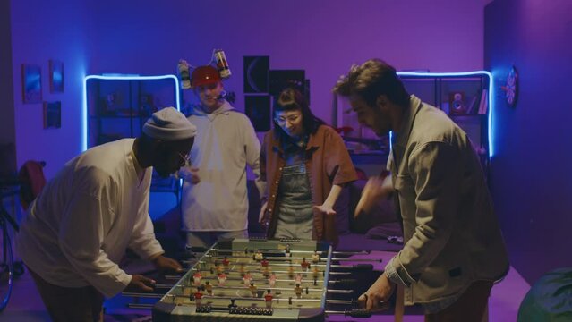 Medium shot of young multiethnic students having fun at home party, African American and Middle Eastern guys playing table football, and Caucasian couple with beer hat dancing to music