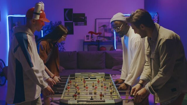 Medium shot of multiethnic young people in casual outfits playing table football in two teams at house party in blue neon lights, African American and Arab guy winning and doing high-fives