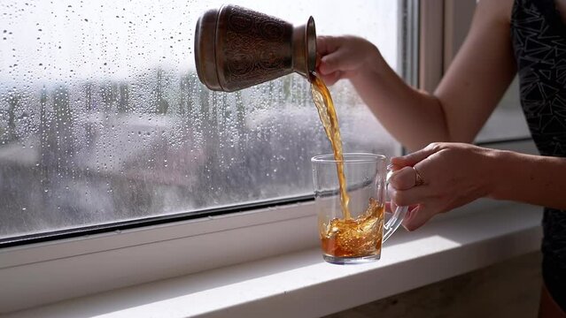Girl Pouring Prepared Turkish Coffee into a Cup while Standing by Window. A hot drink, tea is poured into a transparent glass on background of a sunny rainy day. Sunlight. Drink coffee in the morning.