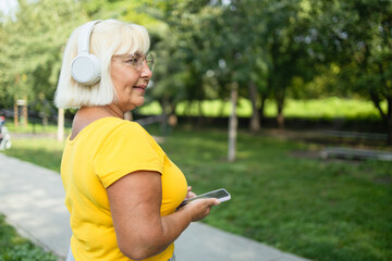 Cheerful caucasian mature woman wife using smart phone cellphone, choosing sound track online while listening to the music in headphones in park. High quality photo