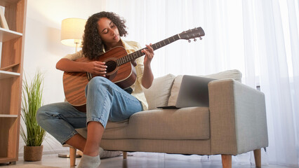Obraz na płótnie Canvas Song learning. Guitar playing. Music hobby. Focused woman musician practicing chords on acoustic instrument sitting couch with laptop home room.
