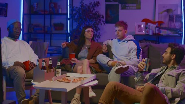 Medium full shot of young Caucasian girl, African American and Arab guys sitting together on couch at home, eating pizza, drinking beer, watching movie or TV show, and discussing events on screen