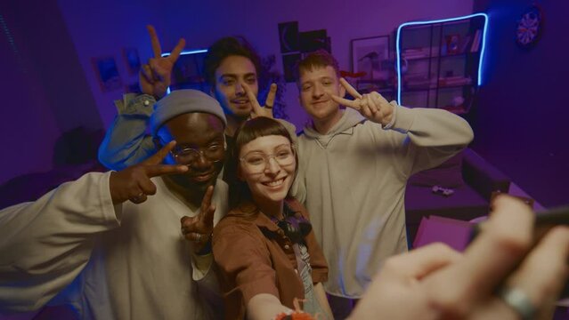 Medium high-angle shot of group of young diverse friends standing in middle of room in blue neon light at home party, shooting selfie photos on smartphone, making funny faces and horns gestures