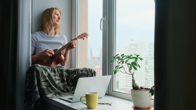 Middle shot relaxed adult blonde woman playing ukulele sitting on windowsill at home. Beautiful female musician enjoying hobby and freedom of improvisation.