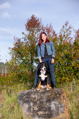 Naklejka na ściany i meble Young beautiful redhead woman standing with her big Bernese Mountain dog, on a rock in a sunny afternoon.