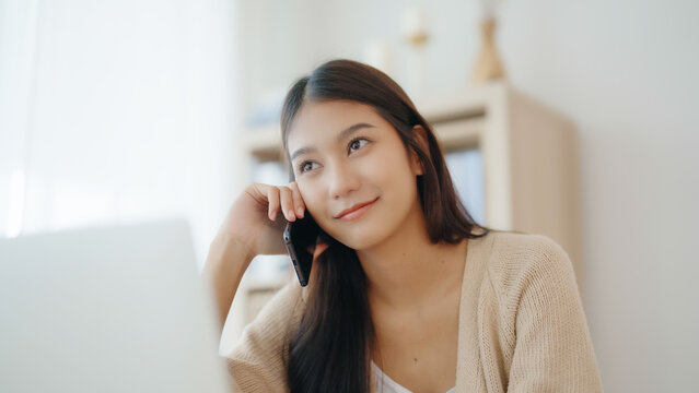 Young asian woman talking mobile smartphone while working on computer laptop on desk in living room at home