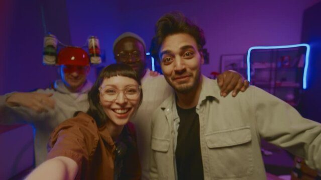 Medium selfie shot of four young multiracial friends shooting video on smartphone for story in social networks at student home party in neon light, fooling around, making faces, smiling and talking