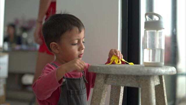 Little latin hispanic boy playing with a yellow broken plastic frog toy on a wooden stool