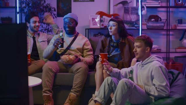 Full shot of young Caucasian woman and three diverse male friends sitting on couch at home on Friday night, drinking beer with crisps, watching sport match or TV show, pointing at screen and arguing