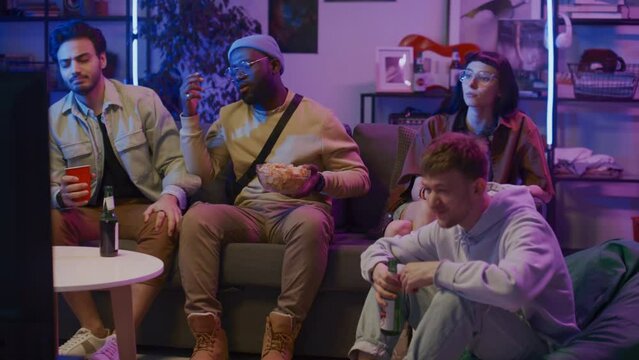 Full shot of young woman and three diverse male friends sitting on couch at home on Friday night, drinking beer with crisps, anxiously watching sport match on TV and shaking head in disappointment