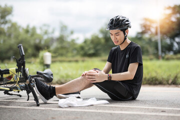 An injured young Asian male cyclist in sportswear and a bike helmet fell off the bike while biking along country roads. knee pain, knee bleeding, injured knee