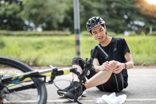 An injured young Asian male cyclist in sportswear and a bike helmet fell off the bike while biking along country roads. knee pain, knee bleeding, injured knee