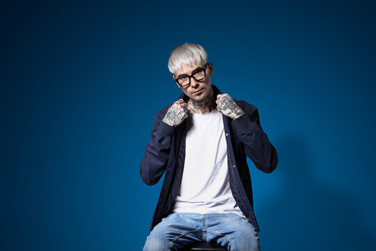 Young tattooed man in eyeglasses sitting on blue background