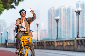 Asian man is riding a bike and using mobile phone to take photo outdoor in the city. 