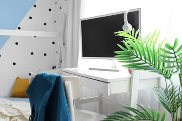 Desk with modern computer and houseplant in interior of children's bedroom