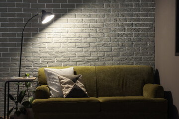 Glowing lamp and cozy green sofa near grey brick wall