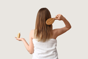 Young woman using cosmetic oil and brush for hair treatment on light background, back view