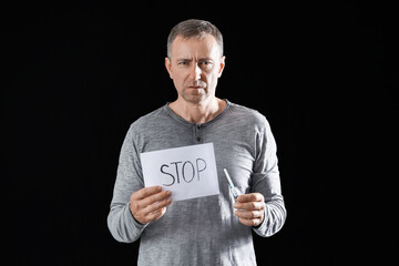 Mature drug addict holding paper with word STOP and syringe on black background