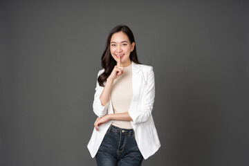 Fototapeta na wymiar Asian female executive with long hair stand and pose Raise your hand and index finger to your mouth and smile. Wearing a white suit, jeans, making eye contact and standing in a gray screened studio.