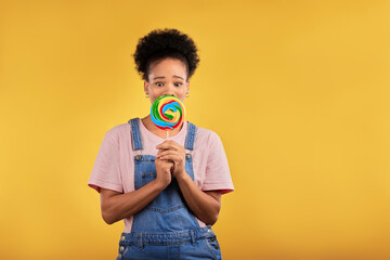 Black woman, portrait with candy or lollipop in studio on yellow background and eating sweets,...